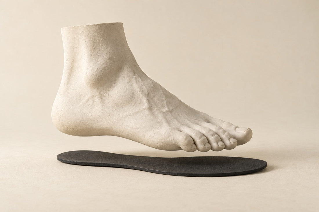 Ivory plaster foot hovering above a thin charcoal sole on a warm ivory background.